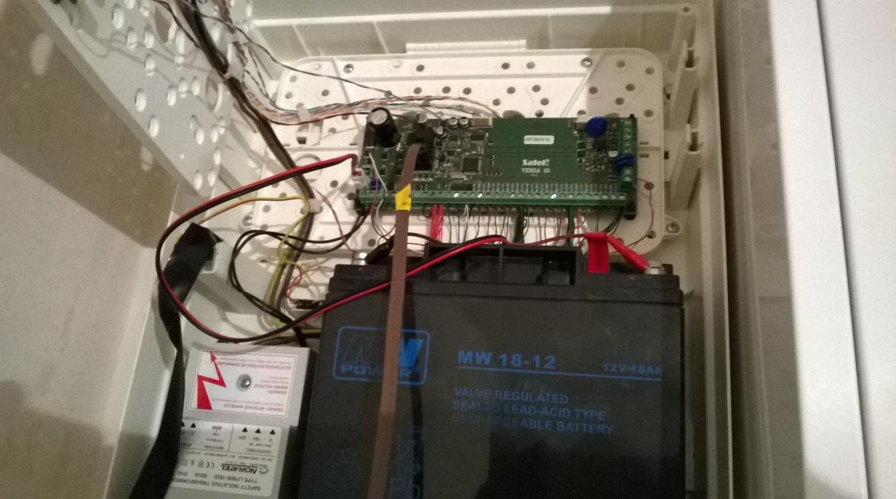 Wnętrze białej skrzynki z elektroniką, akumulatorem MW 18-12 i transformatorem Noratel, widoczne przewody i płytka drukowana Safet VEDNA 15.