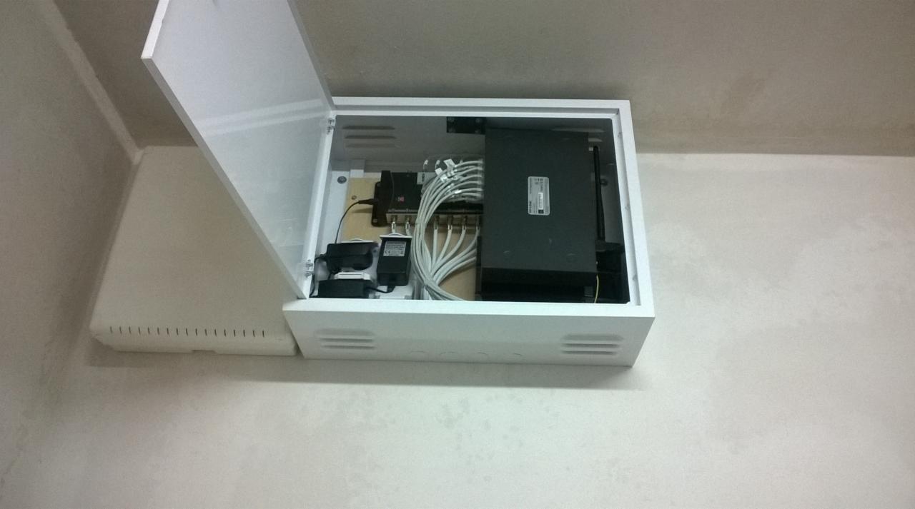 Otwarta, biała skrzynka montażowa na ścianie, zawierająca router, zasilacze i okablowanie strukturalne, widok od dołu.