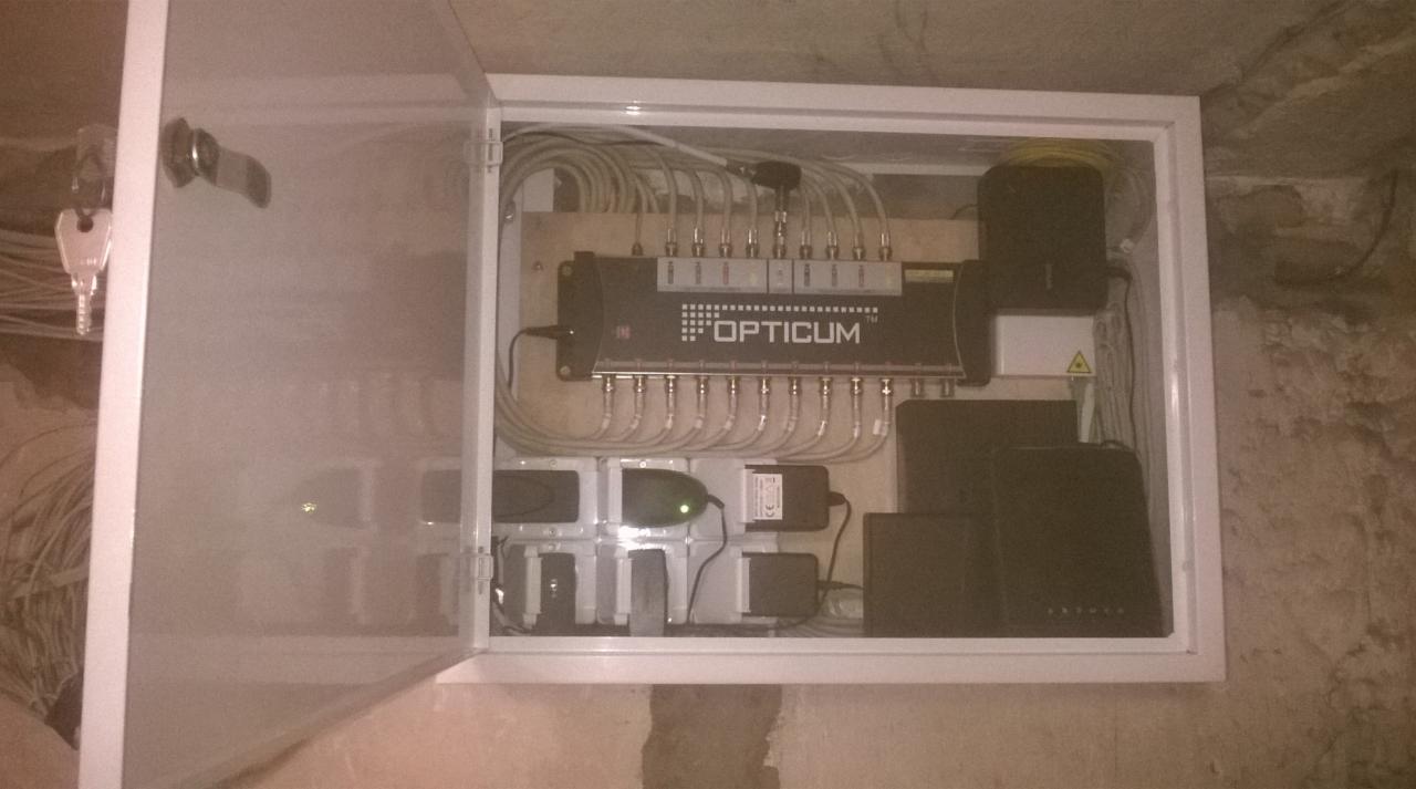 Otwarta skrzynka z rozdzielnicą sygnału OPTICUM, kablami i zasilaczami, zamontowana na ścianie z surowym tynkiem. Klucze wiszą na drzwiczkach.