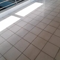 Błyszcząca, szara podłoga z płytek w hali basenowej, oświetlona promieniami słońca. Widoczna balustrada i fragment basenu w tle. Czystość i porządek.