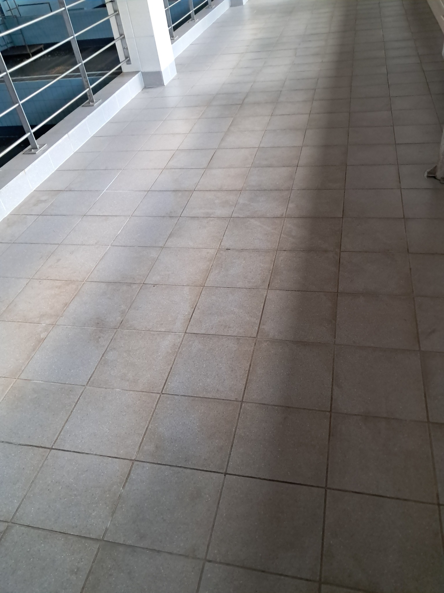 Korytarz wyłożony szarymi, kwadratowymi płytkami. Widoczna balustrada ze stali nierdzewnej i fragment ściany. Podłoga z widocznymi śladami użytkowania.