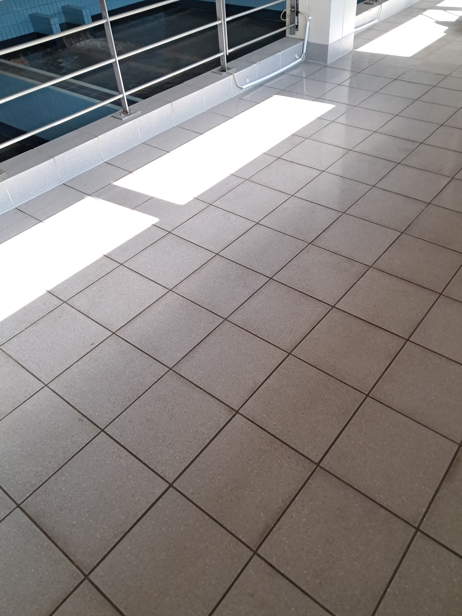 Błyszcząca, szara podłoga z płytek w hali basenowej, oświetlona promieniami słońca. Widoczna balustrada i fragment basenu w tle. Czystość i porządek.
