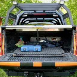 Pomarańczowy pickup z otwartą przestrzenią ładunkową, wyposażony w metalową konstrukcję nad kabiną z dodatkowym oświetleniem LED, widoczne narzędzia i skrzynki w przestrzeni ładunkowej na tle zieleni.