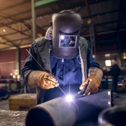 Pracownik w masce spawalniczej łączy dwa metalowe elementy rury podczas pracy w warsztacie, widoczne iskry i intensywne światło łuku elektrycznego, w tle zarys innych maszyn i postaci.