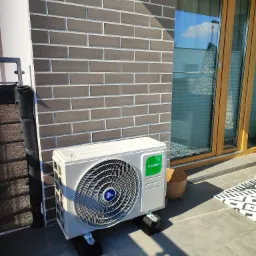 Zewnętrzna jednostka klimatyzacji Kaisai na szarym tarasie, przy ścianie z cegły. Widoczne okno z odbiciem nieba i element dekoracyjny.