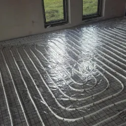 Białe rury ogrzewania podłogowego ułożone na srebrnej folii i siatce w surowym pomieszczeniu, widok z góry na system instalacji przed zalaniem wylewką.