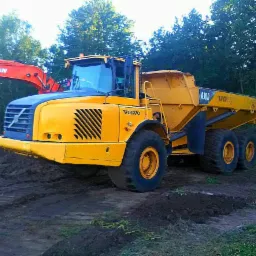 Żółta wywrotka Volvo A30D stoi na ubitej ziemi, z pomarańczową koparką Doosan widoczną w tle, otoczona drzewami.