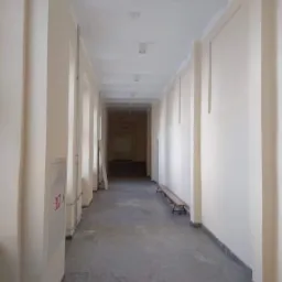 Długi, pusty korytarz z beżowymi ścianami i szarą podłogą z lastryko, oświetlony lampami sufitowymi. Widoczny fragment ławki pod ścianą i skrzynka przeciwpożarowa. Na ścianach widoczne panele...