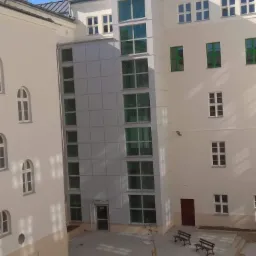 Wewnętrzny dziedziniec budynku z panelami elewacyjnymi, widok z góry, z ławkami i młodym drzewem.