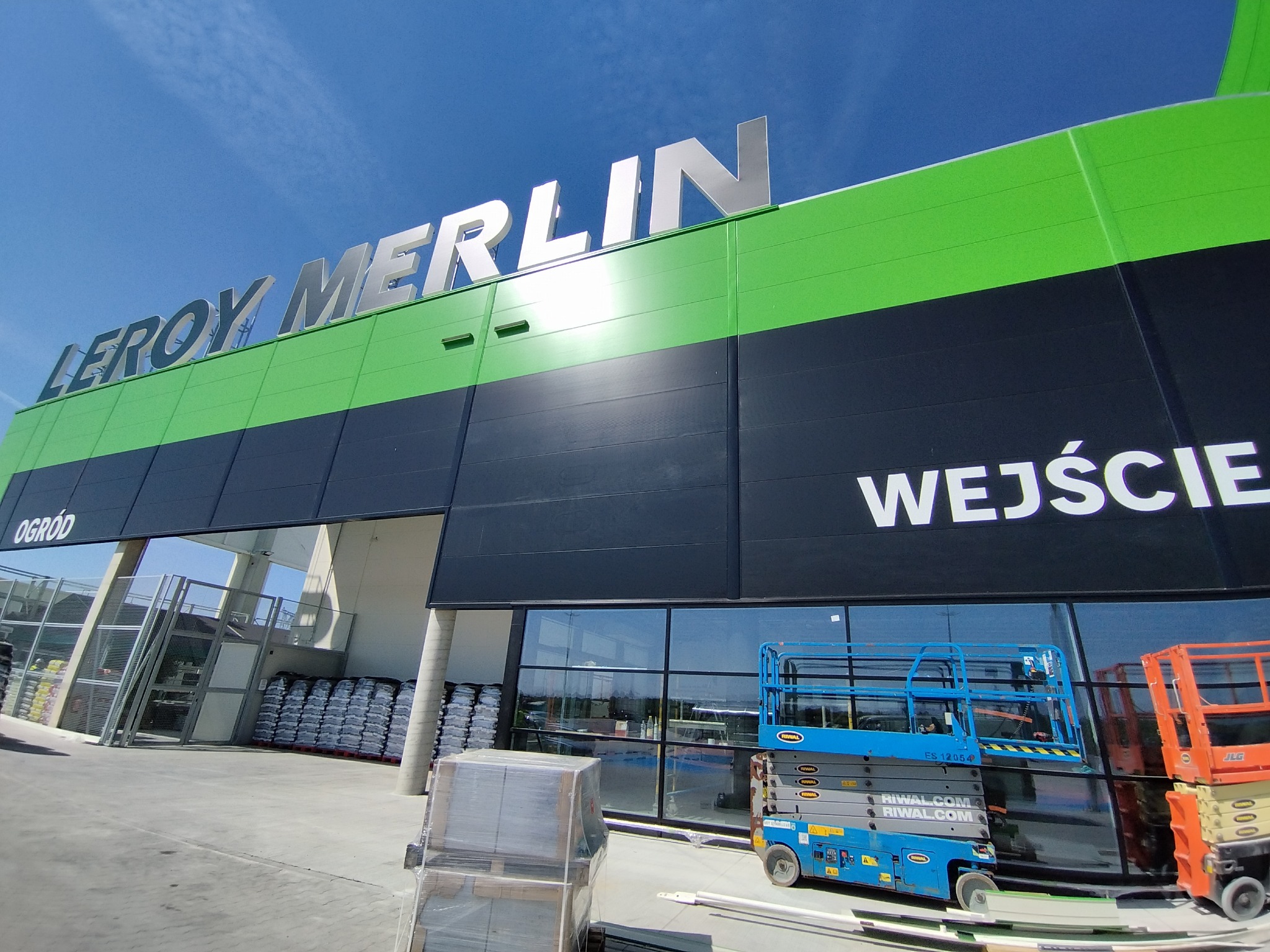 Widok na budynek sklepu budowlanego Leroy Merlin z napisem 'WEJŚCIE', z widocznymi podnośnikami koszowymi i materiałami budowlanymi na zewnątrz.