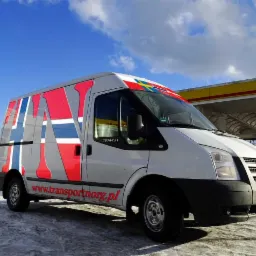 Biały bus marki Ford Transit z kolorowym wzorem przypominającym flagi skandynawskie i adresem strony internetowej transportnorg.pl, zaparkowany na śniegu przed stacją benzynową w słoneczny dzień.