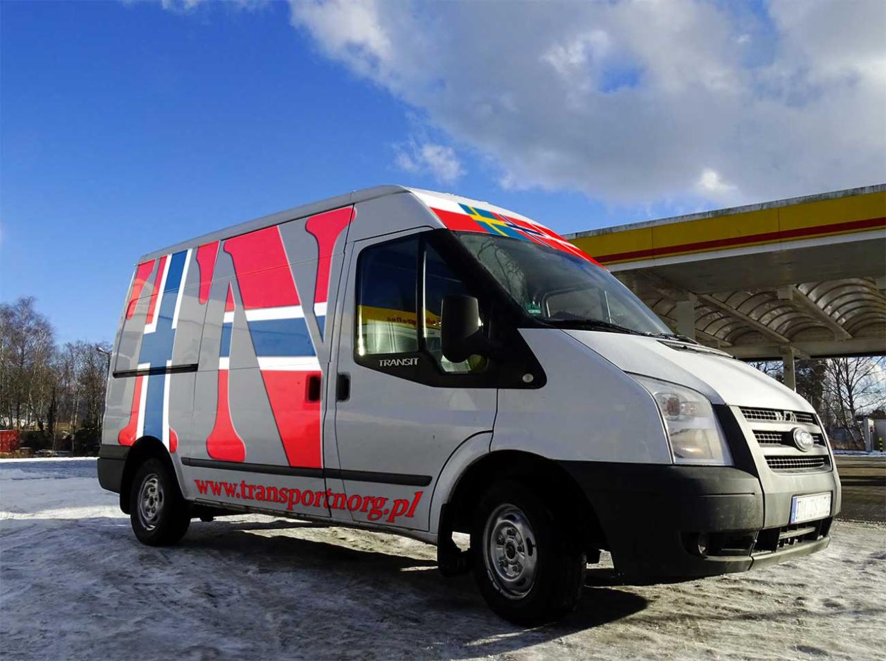 Biały bus marki Ford Transit z kolorowym wzorem przypominającym flagi skandynawskie i adresem strony internetowej transportnorg.pl, zaparkowany na śniegu przed stacją benzynową w słoneczny dzień.