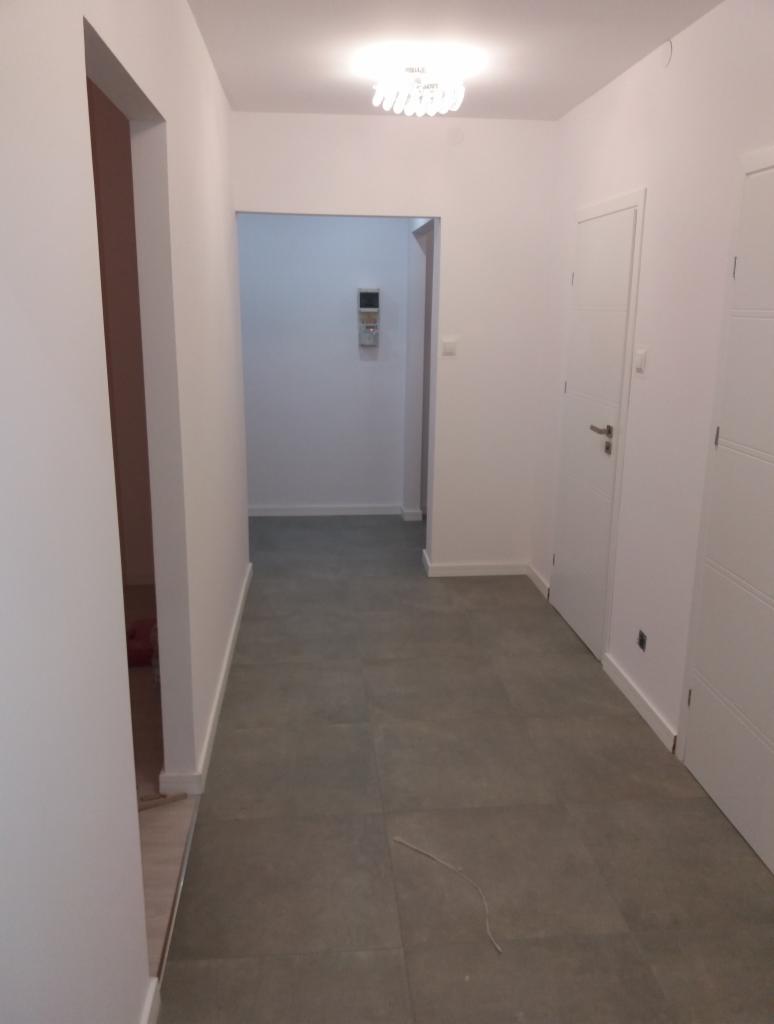 Korytarz w Katowicach z szarymi płytkami podłogowymi, białymi ścianami i drzwiami. Widoczny fragment otwartego przejścia do innego pomieszczenia z regulatorem temperatury na ścianie.