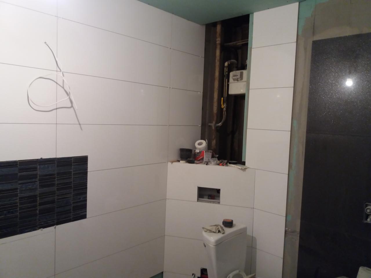 Układanie białych, prostokątnych płytek na ścianie w łazience, widoczny fragment instalacji gazowej w otworze, czarne płytki dekoracyjne oraz wiszące przewody elektryczne.