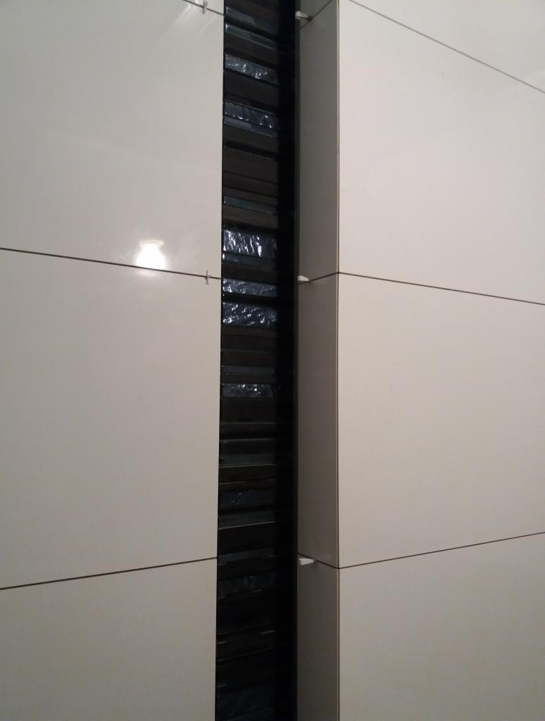 Ściana z jasnych, błyszczących paneli z wąskim, pionowym pasem ciemnych, strukturalnych płytek między nimi, zabezpieczonych folią ochronną.