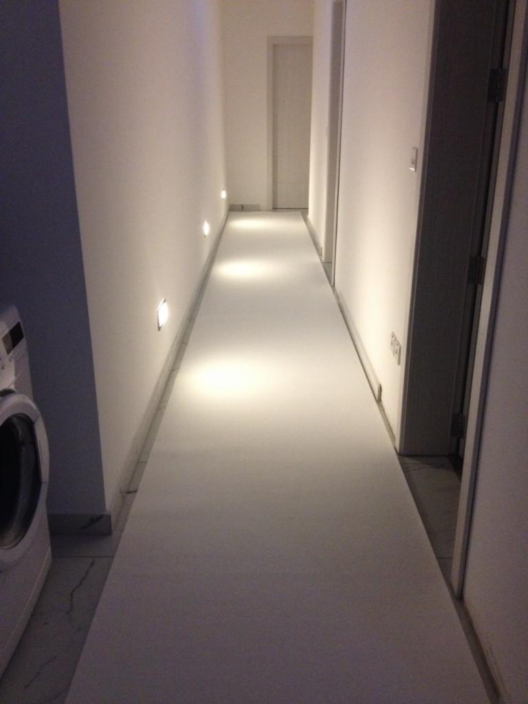 Wąski korytarz z białymi ścianami i podłogą, oświetlony punktowymi lampami w ścianie, widoczna pralka po lewej stronie.