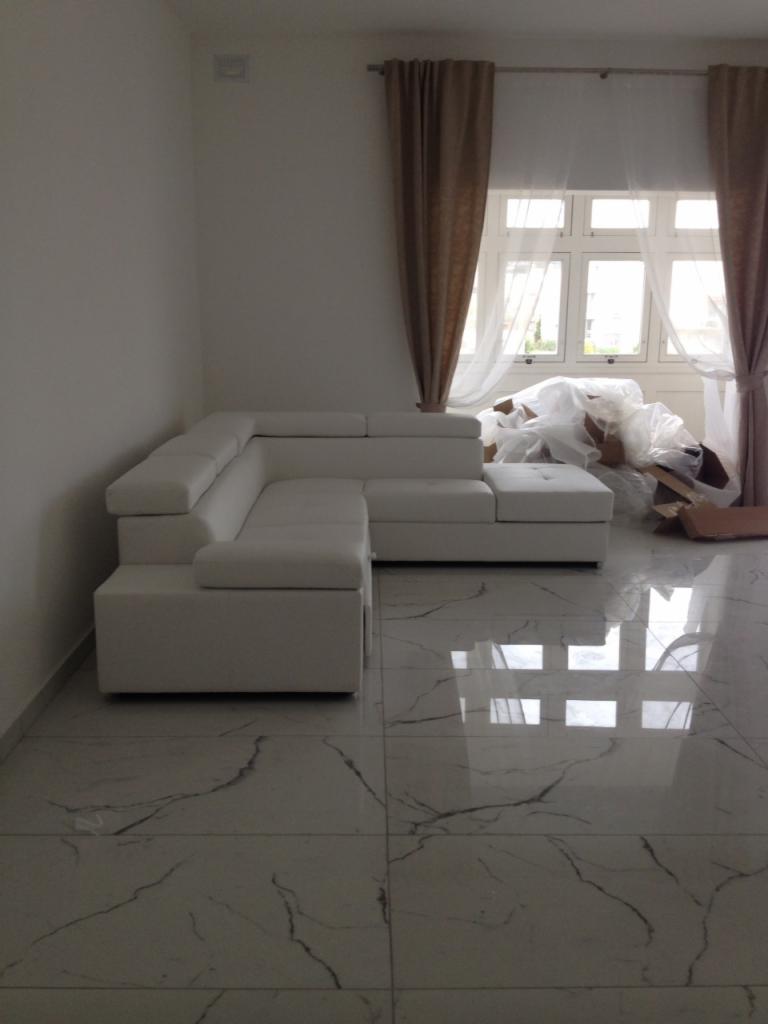 Jasny narożnik modułowy z ekoskóry na błyszczącej podłodze imitującej marmur, z widocznymi śladami po rozpakowywaniu mebli przy oknie z brązowymi zasłonami.