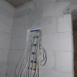 Białe przewody elektryczne ułożone w peszlach na surowej ścianie z betonu komórkowego, prowadzące do puszki rozdzielczej.