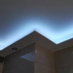 Podwieszany sufit z oświetleniem LED w narożniku łazienki wyłożonej beżowymi płytkami, widoczny okrągły reflektor i kratka wentylacyjna.