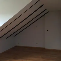 Poddasze z drewnianą podłogą i skośnym sufitem z widocznymi czarnymi belkami konstrukcyjnymi, przygotowane do dalszych prac wykończeniowych.