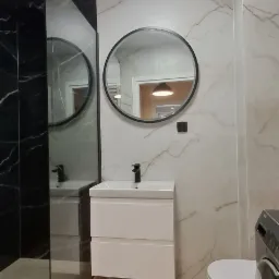 Nowoczesna łazienka z kabiną prysznicową typu walk-in z czarnymi elementami, okrągłym lustrem nad białą szafką z umywalką, marmurowe płytki na ścianach i beżowe na podłodze, widoczna pralka.