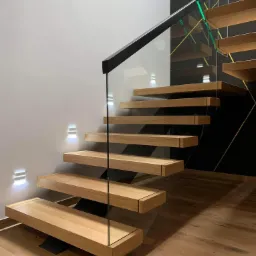 Nowoczesne, drewniane schody wiszące z balustradą ze szkła i czarnymi elementami mocującymi, oświetlone kinkietami na ścianie, z widoczną podłogą z desek.