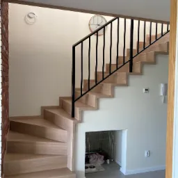 Jasne drewniane schody z czarną metalową balustradą w nowoczesnym wnętrzu, pod schodami klatka dla psa z miskami i legowiskiem.