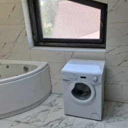 Biała łazienka z wanną jacuzzi, pralką Candy Aquamatic i podwieszaną toaletą, wykończona płytkami imitującymi marmur, z oknem w ukośnej wnęce.