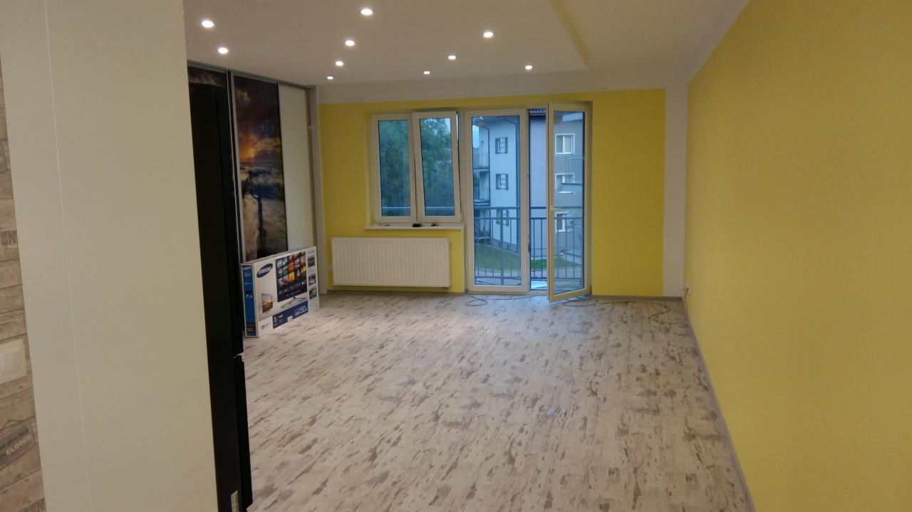 Pomalowane na żółto wnętrze pokoju z panelami podłogowymi imitującymi drewno, z oknem balkonowym i grzejnikiem oraz przesuwane drzwi z grafiką krajobrazu.