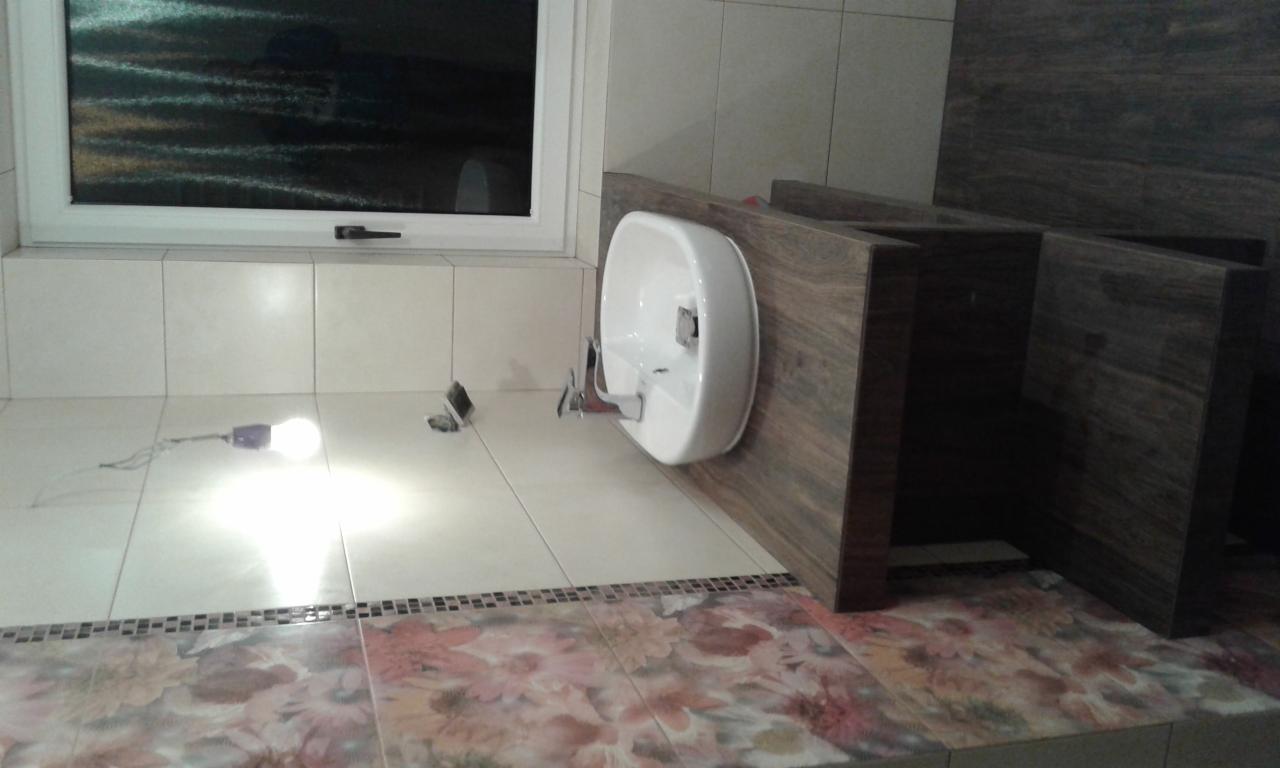 Niedokończony montaż umywalki na drewnianej szafce w łazience w Czechowicach-Dziedzicach, widoczna żarówka leżąca na podłodze i płytki z motywem kwiatowym.