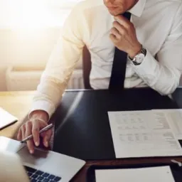 Mężczyzna w białej koszuli i krawacie siedzi przy biurku z laptopem, dokumentami i kawą, zamyślony, trzymając dłoń przy brodzie.