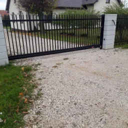 Czarna, przesuwna brama wjazdowa z pionowymi prętami zamontowana pomiędzy szarymi słupkami z bloczków betonowych, na żwirowym podjeździe, widok z zewnątrz posesji.