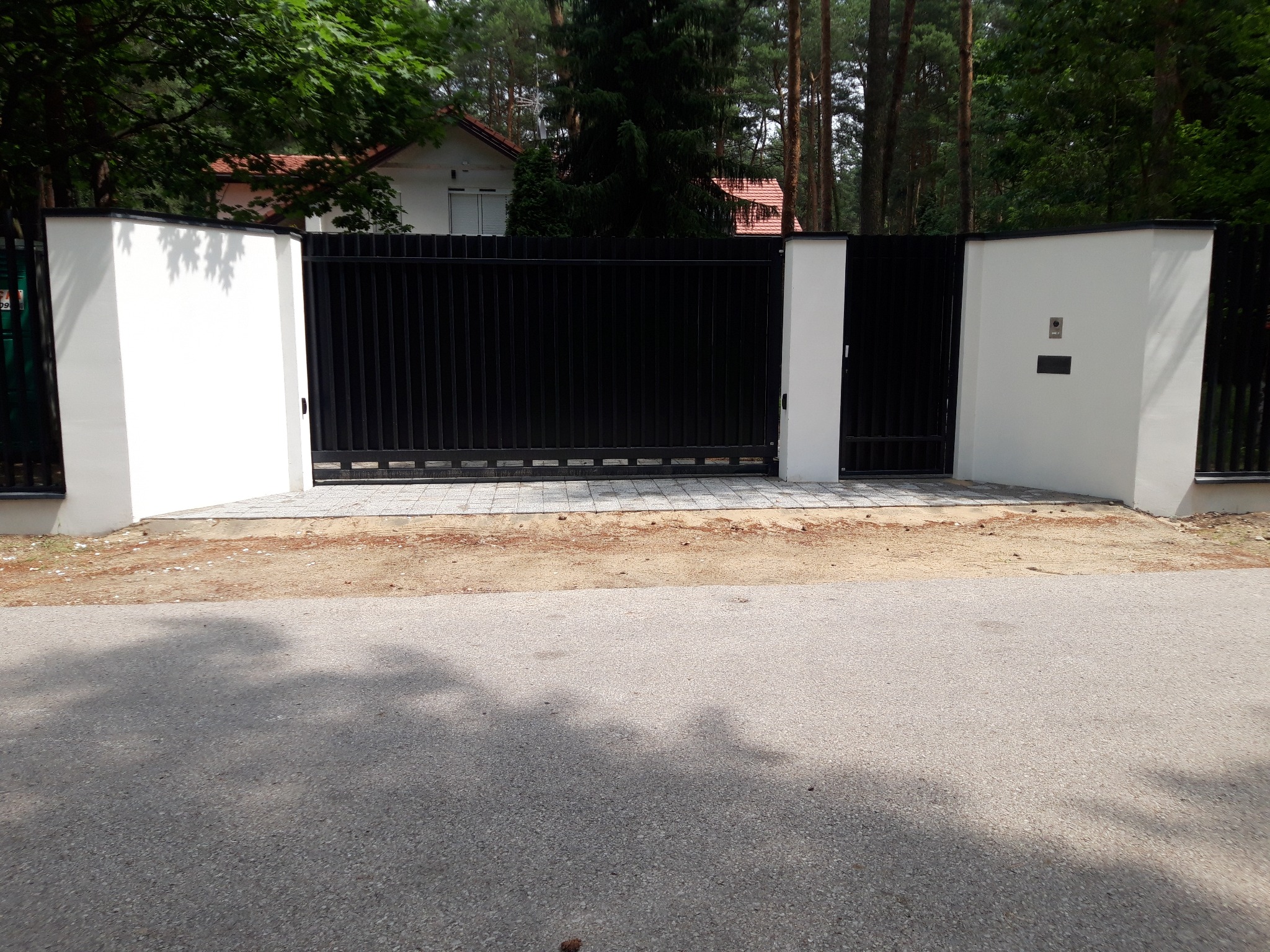 Nowoczesna, czarna brama wjazdowa z pionowymi panelami, osadzona między białymi, masywnymi słupkami, prowadząca do posesji z widocznym fragmentem domu z czerwoną dachówką w tle, ułożona kostka...