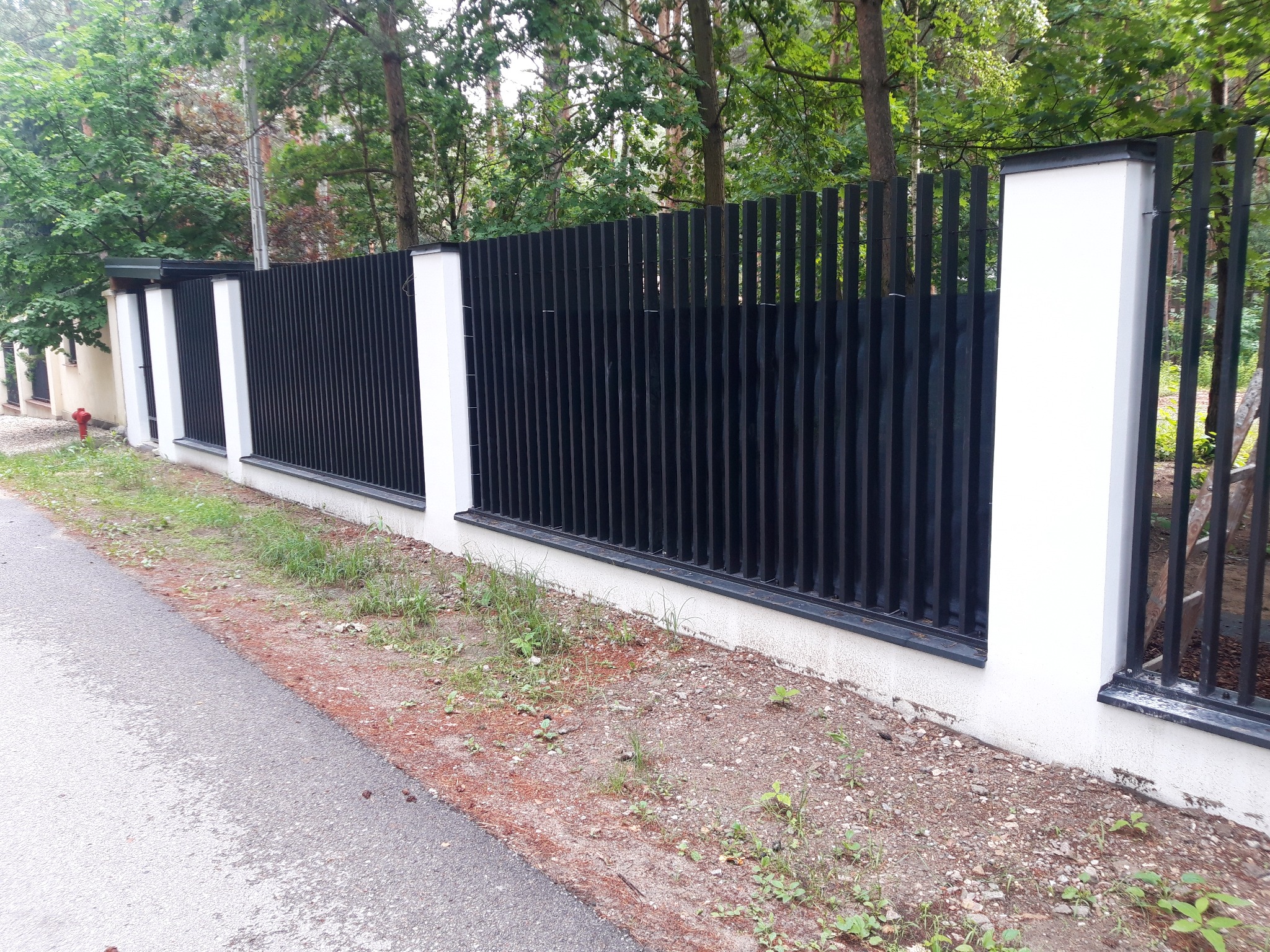 Nowoczesne ogrodzenie z czarnych, pionowych elementów metalowych i białych, kwadratowych słupków, widok z zewnątrz posesji.