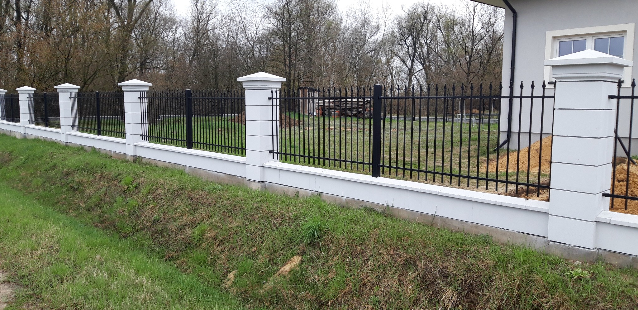 Nowoczesne ogrodzenie z czarnych, pionowych prętów i białych, segmentowych słupków, z widoczną konstrukcją budynku w tle.