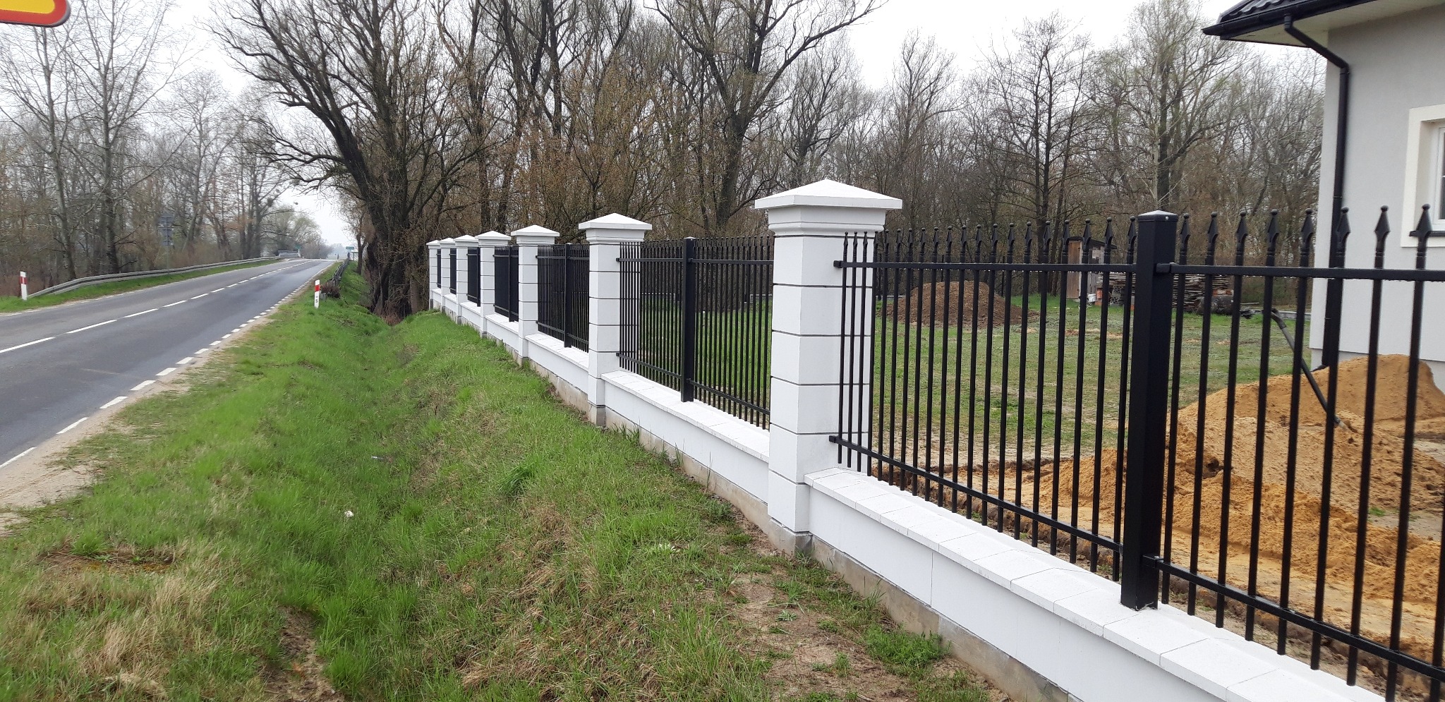 Nowoczesne ogrodzenie z czarnych prętów i białych słupków z dekoracyjnymi kapitelami, oddzielające posesję od drogi z drzewami.