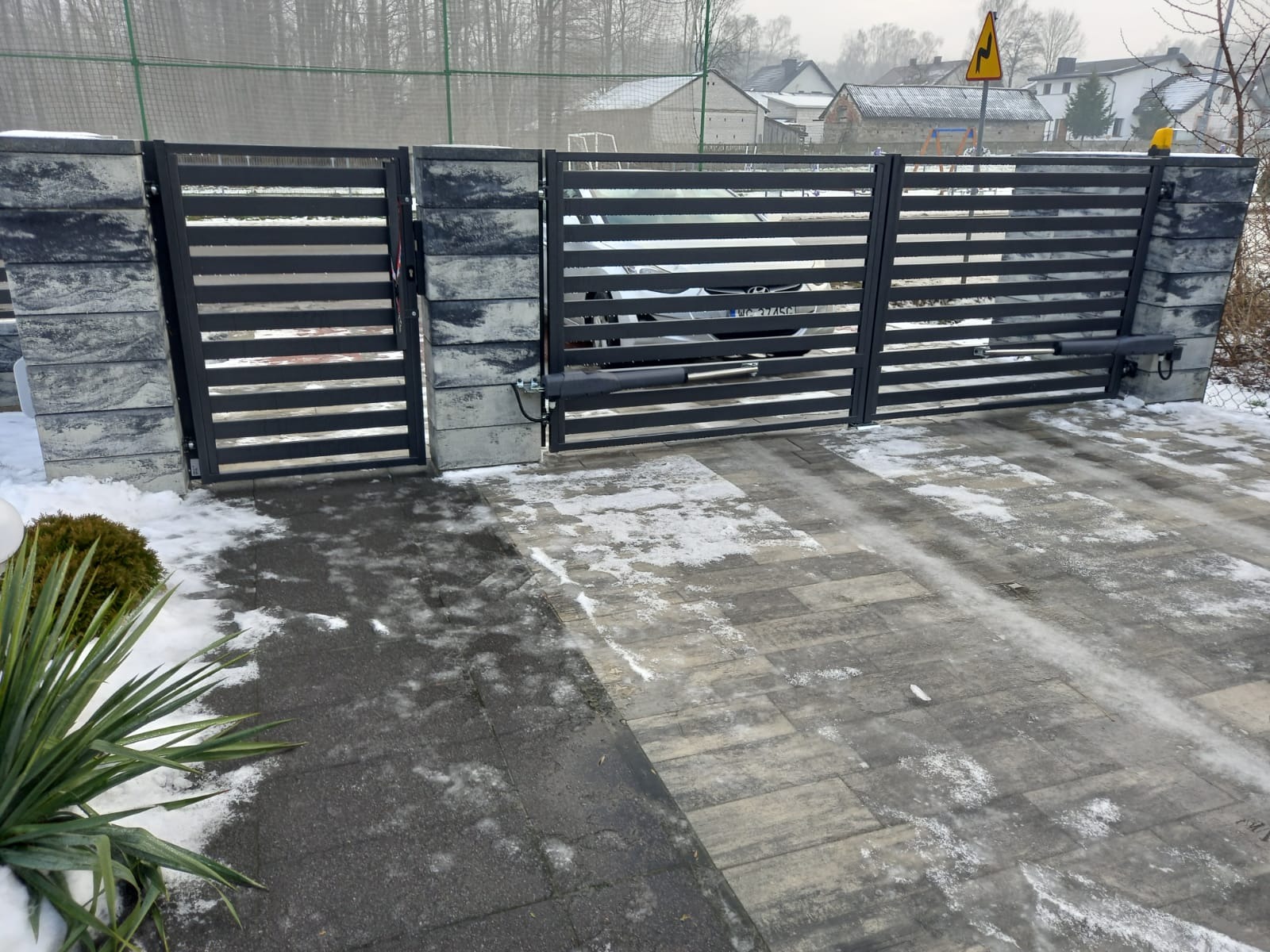 Nowoczesna brama wjazdowa z poziomymi profilami, zamontowana na słupkach z szarego betonu architektonicznego, z automatyką widoczną po prawej stronie, na podjeździe częściowo pokrytym śniegiem.