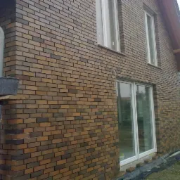 Zewnętrzna ściana domu z brązowej cegły klinkierowej, widoczne okna i drzwi balkonowe z białymi ramami, na pierwszym planie rusztowanie z wiadrami, w tle drugi budynek i betoniarka.