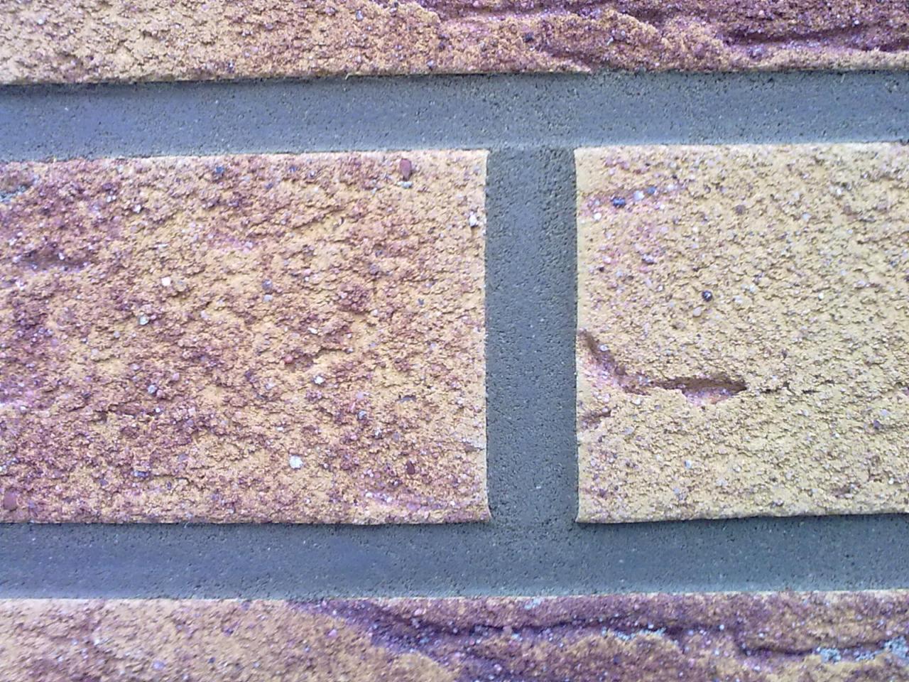 Fragment murowanej ściany z cegły klinkierowej w odcieniach żółci i fioletu, z widocznymi szarymi spoinami.