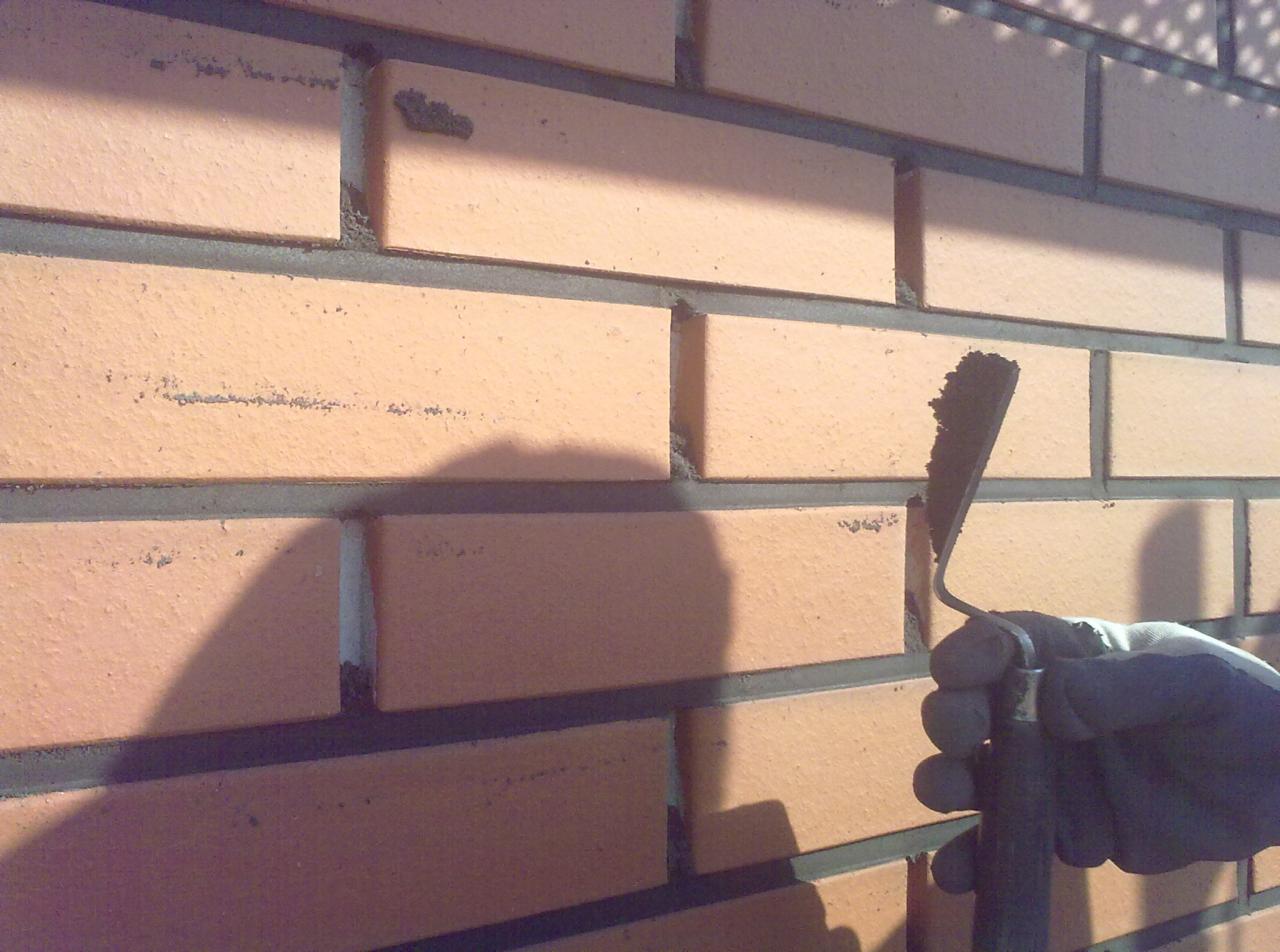 Murowana ściana z cegły klinkierowej w trakcie budowy, widoczna dłoń w rękawicy trzymająca kielnię z zaprawą, cień dłoni na murze.