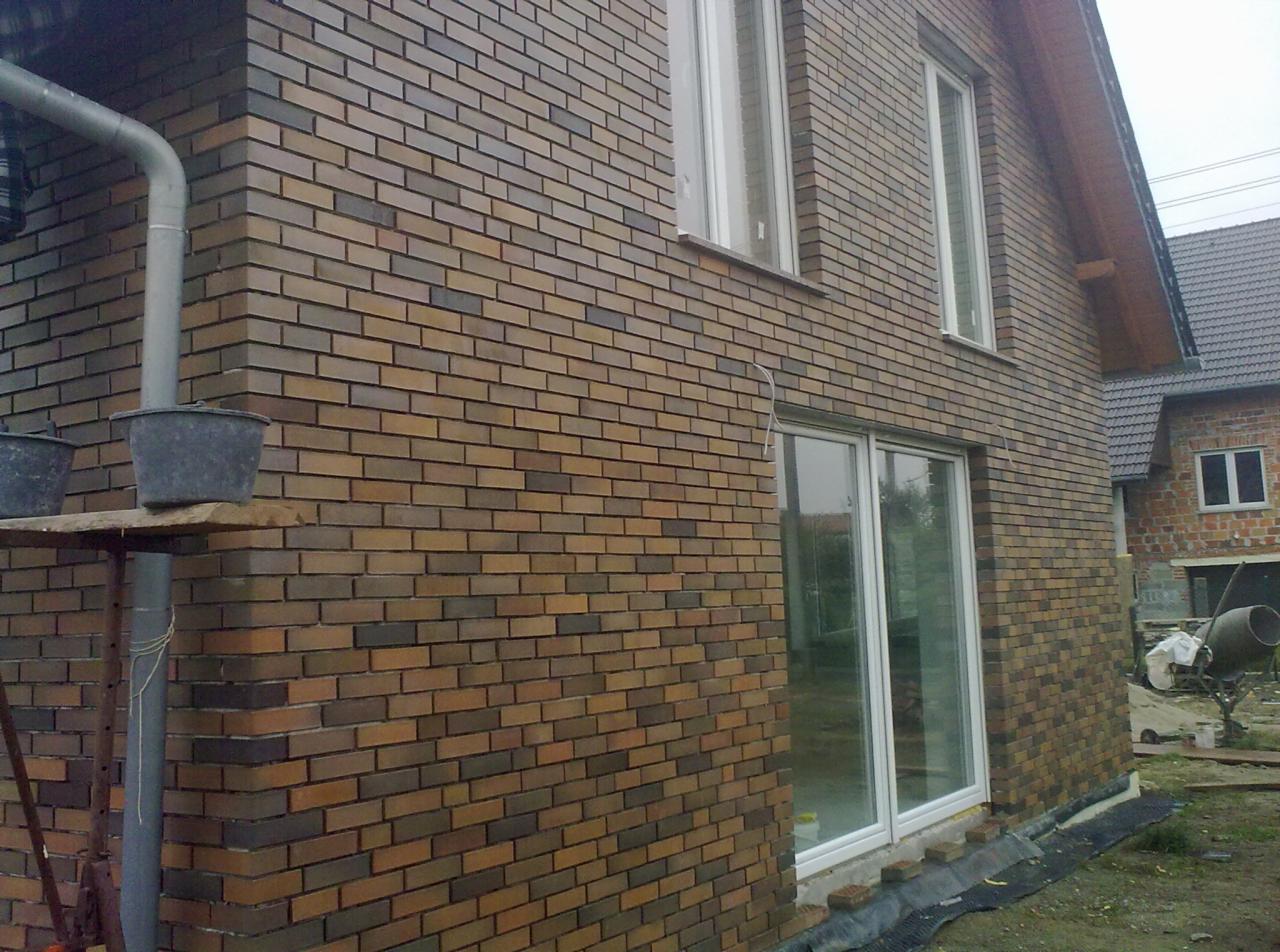 Zewnętrzna ściana domu z brązowej cegły klinkierowej, widoczne okna i drzwi balkonowe z białymi ramami, na pierwszym planie rusztowanie z wiadrami, w tle drugi budynek i betoniarka.