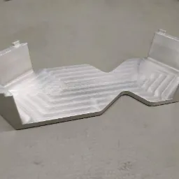 Precyzyjnie obrobiony, aluminiowy element z wyraźnymi śladami frezowania, leżący na szarym podłożu. Widoczne symetryczne kształty i kąty proste.