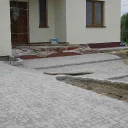 Fragment posesji w Górze Kalwarii w trakcie prac brukarskich: ułożona szara kostka na podjeździe i schodach wejściowych, widoczne przygotowane obrzeża i materiały budowlane.