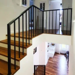 Schody drewniane z czarną, metalową balustradą, widok z góry na otwartą przestrzeń z kuchnią w tle.