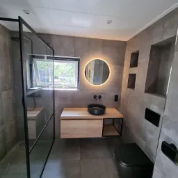 Nowoczesna łazienka z szarymi płytkami, czarną armaturą i okrągłym lustrem z podświetleniem LED, kabina prysznicowa walk-in z czarną ramą, wisząca szafka z drewnianym blatem i czarną umywalką.