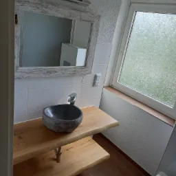 Umywalka z kamienia w kształcie misy na drewnianym blacie w łazience z oknem, lustro w białej ramie z efektem postarzenia, białe płytki na ścianie, podłoga z paneli imitujących drewno.