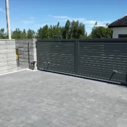 Automatyczna brama wjazdowa z poziomymi panelami, szara kostka brukowa na podjeździe, fragment ogrodzenia z betonowych bloczków, widoczne siłowniki bramy.