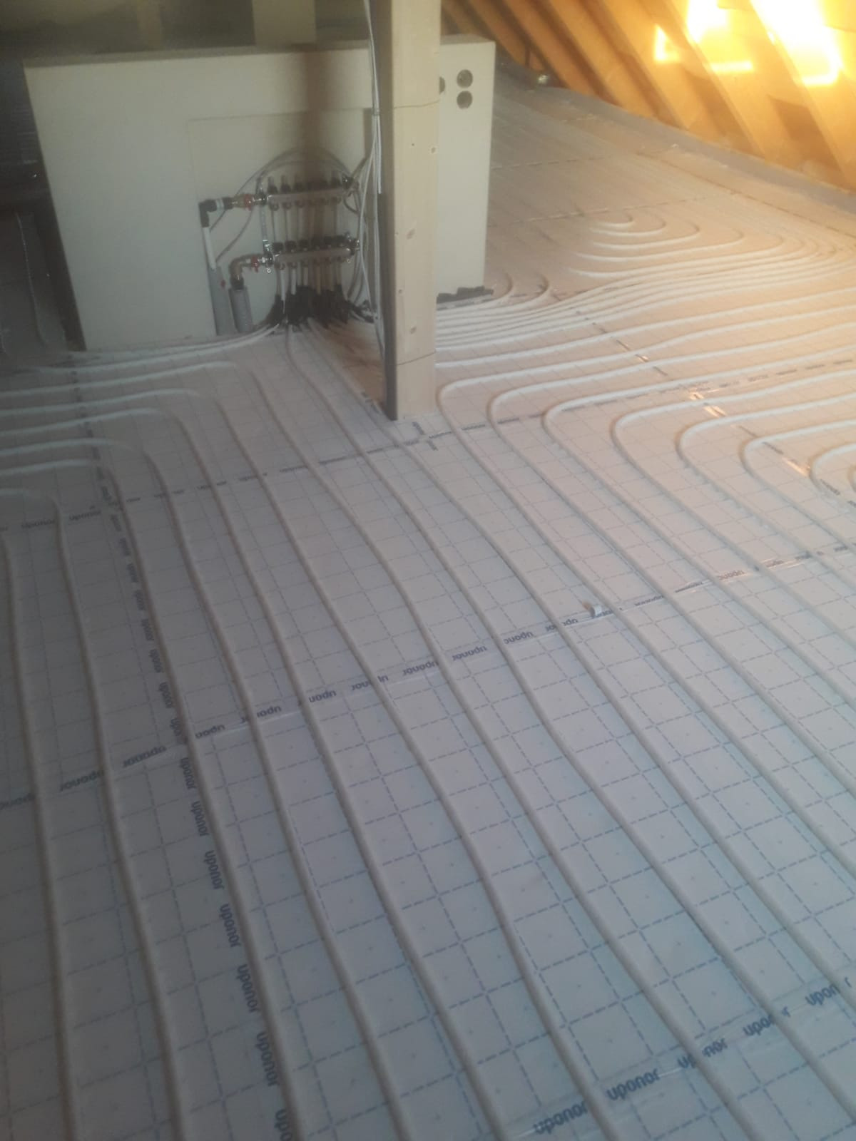 Rozłożone rury ogrzewania podłogowego Uponor na izolacji z widocznym rozdzielaczem i belkami stropowymi w tle.