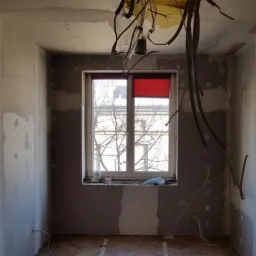 Wnętrze pokoju w trakcie remontu: ściany przygotowane do malowania z widocznymi śladami szpachlowania, odsłonięte przewody elektryczne zwisające z sufitu, okno z częściowo zasłoniętą roletą...