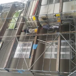 Dwóch pracowników na rusztowaniu podczas nakładania szarego tynku na elewację domu, okna zabezpieczone folią, widoczne narzędzia i żółte wiadra.